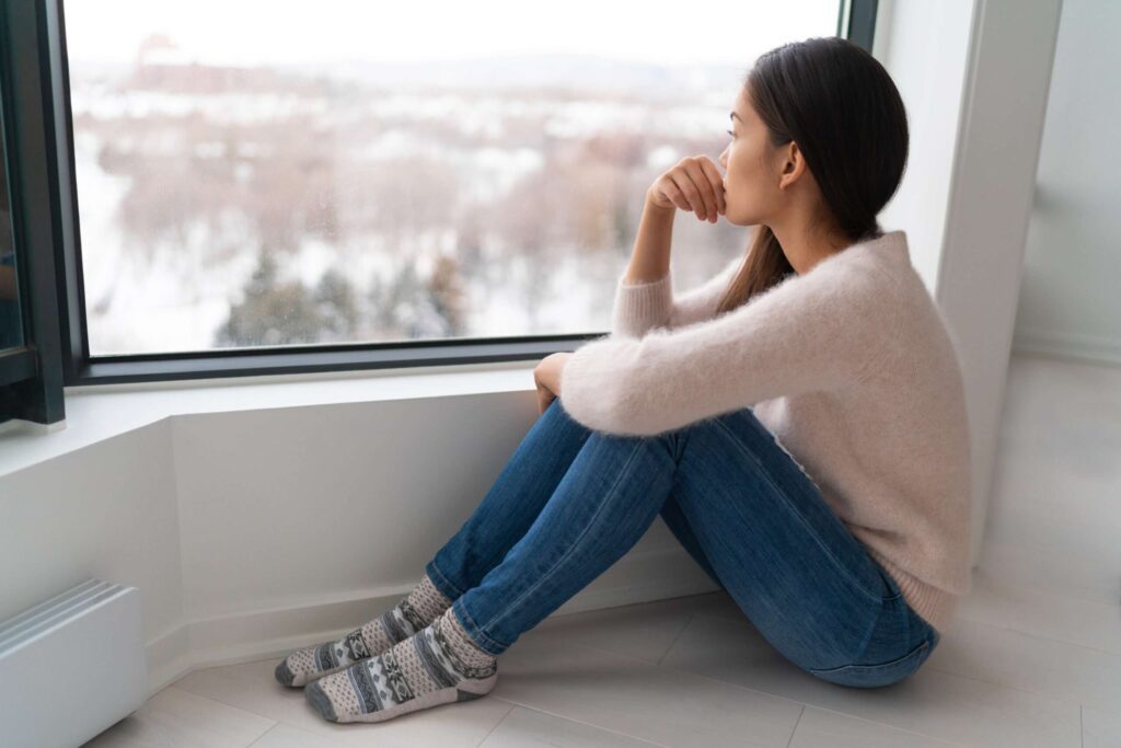 Depressed young girl feeling sad an lonely, anxious looking out the window in winter. Unhappy Asian woman alone, depression at home.